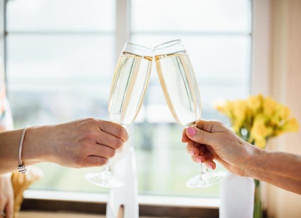 Two hands holding champagne flutes together in a toast near a bright window.