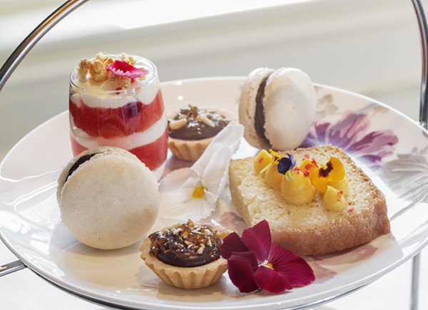 A white plate holding assorted small desserts, including macarons, tartlets, a layered red and white parfait, and a square cake topped with花