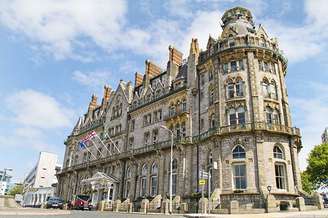 A large historic stone building with a rounded corner tower, flags above the entrance, and a street in front under a blue sky.