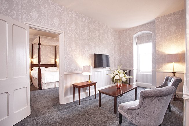 A bright sitting room with patterned wallpaper, two armchairs, a coffee table with flowers, a side table, and a wall-mounted TV, with a four