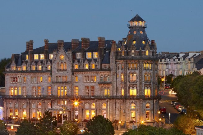 A large historic stone hotel with many lit windows at dusk, seen from outside with trees and a road beside it.