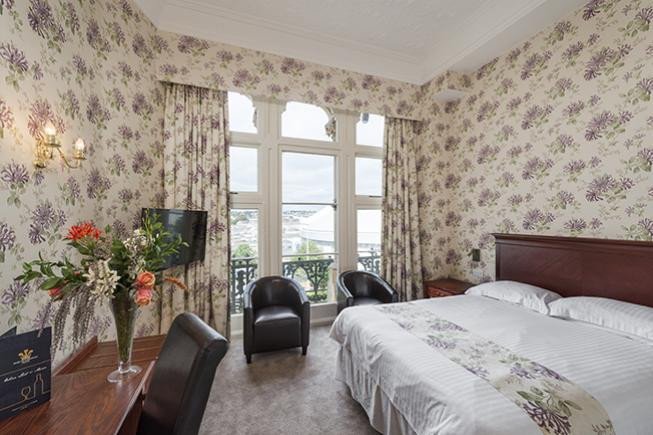 A hotel room with floral wallpaper, a large bed, two black chairs by tall windows, and a balcony view outside.