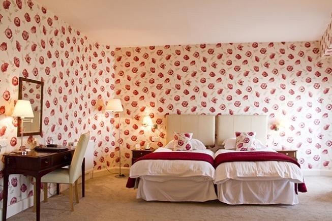 A bedroom with two twin beds, floral wallpaper, bedside lamps, and a desk with a mirror and chair.