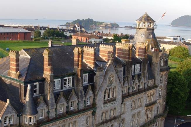 A historic stone building with a slate roof and turret overlooking a coastal harbor with islands in the distance.