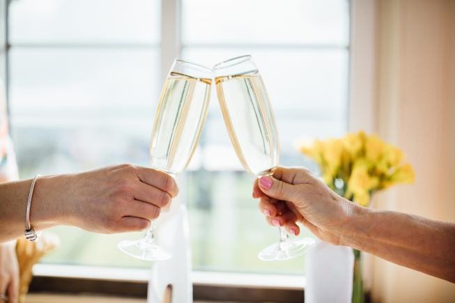 Two people clink champagne flutes together in front of a bright window.