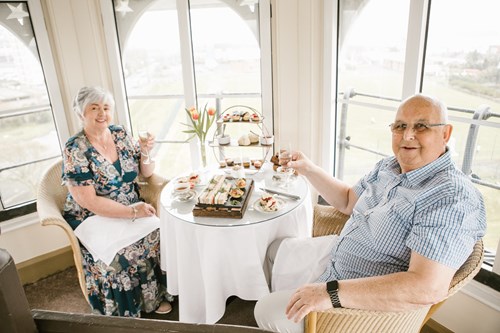 Two people seated at a small table with tea, sandwiches, and pastries, raising glasses in a bright room with large windows.