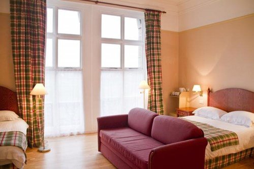 A bedroom with two beds, a maroon sofa, floor lamps, and a large window with plaid curtains.
