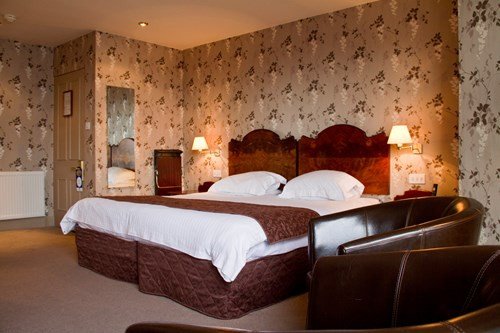 A hotel bedroom with a large bed, floral wallpaper, bedside lamps, and leather chairs in the foreground.