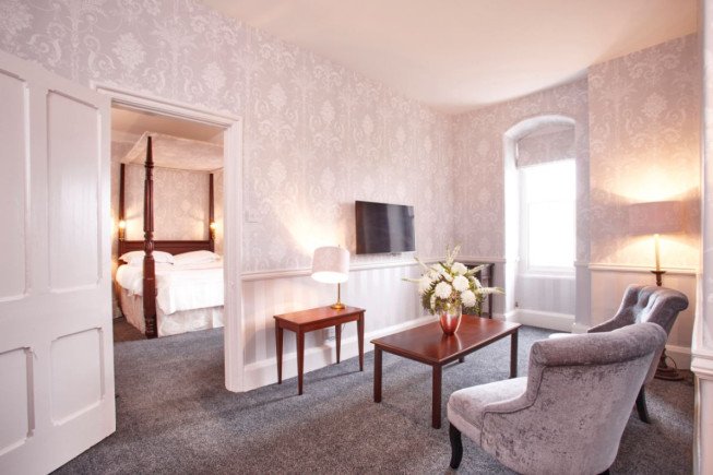 A carpeted room with patterned wallpaper, two upholstered chairs, a coffee table with flowers, a lamp, and a wall-mounted TV, with a bedroom