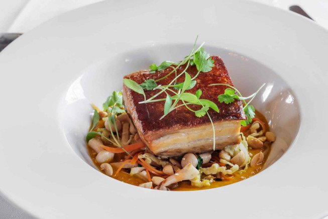 A square piece of seared pork belly garnished with herbs, served in a white bowl over vegetables and beans.