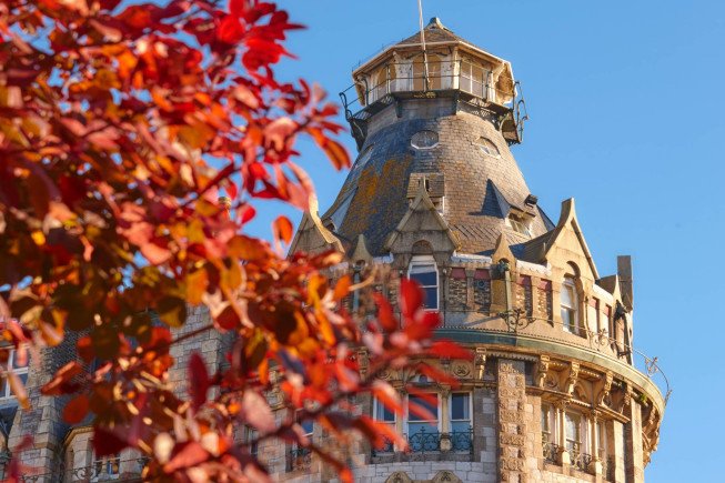 A stone tower with a rounded roof and balconies is seen behind red autumn leaves against a blue sky.