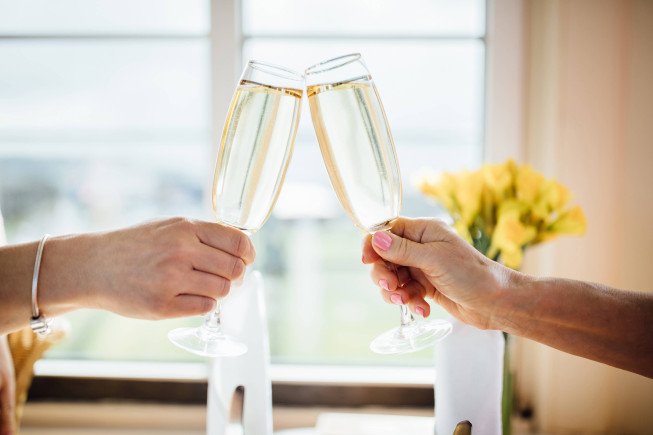 Two hands hold champagne flutes together in a toast near a window.