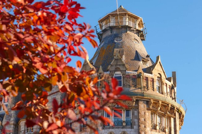 A stone building with a steep, rounded roof and small tower, partly framed by red autumn leaves against a blue sky.