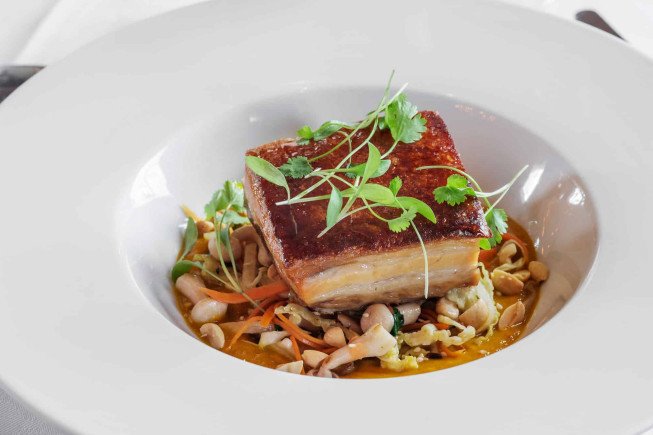 A square piece of seared pork belly topped with herbs, served in a shallow white bowl over mushrooms and vegetables.