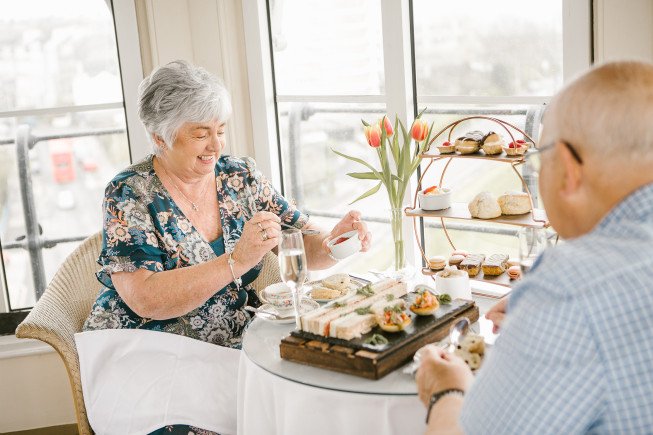 Two people sit at a small table with tea, pastries, and a vase of tulips near a bright window.