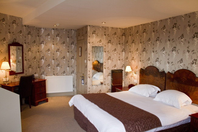 A hotel bedroom with a large bed, bedside lamps, a desk and chair, and floral wallpaper.