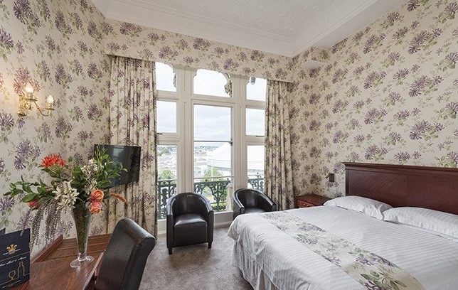 A hotel bedroom with floral wallpaper, a large bed, two black chairs by tall windows, and a desk with flowers.