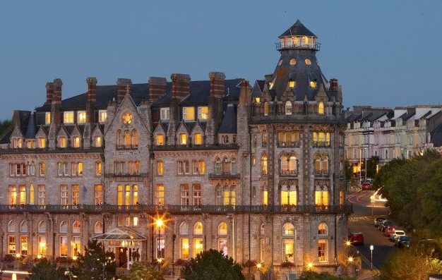 A large historic stone hotel with many lit windows and a tower, seen at dusk against a blue sky.
