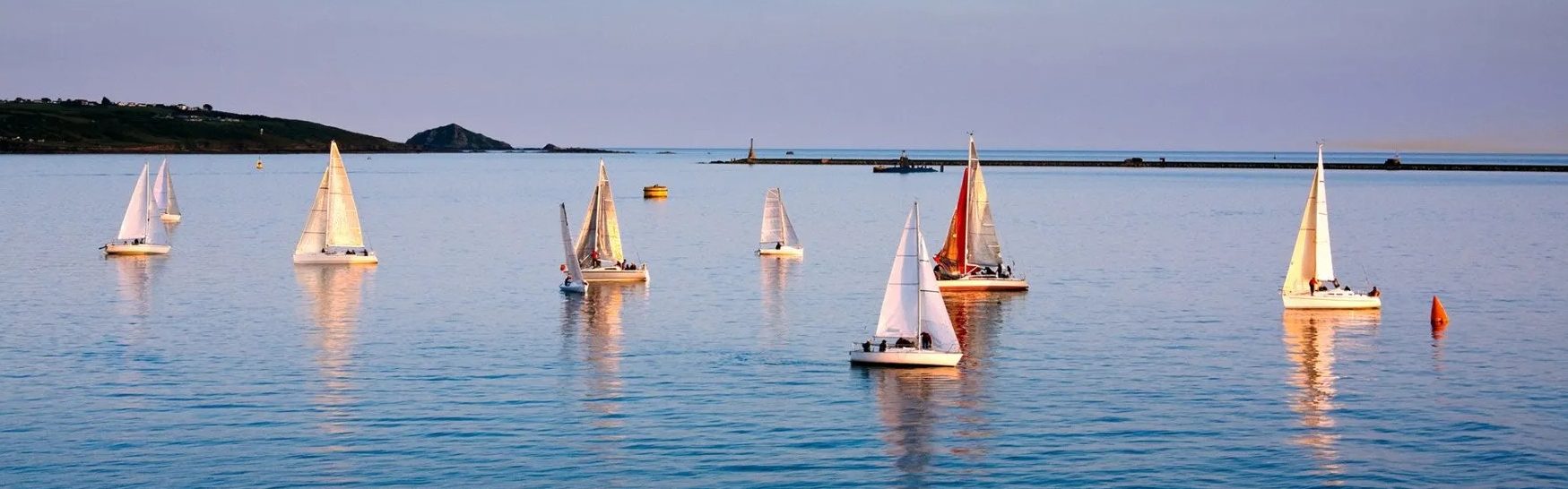 Several sailboats on calm blue water near a harbor breakwater at dusk.
