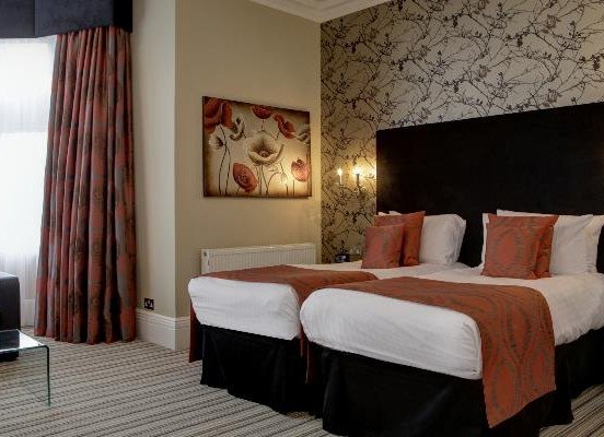 A hotel room with two single beds dressed in white linens and orange runners, patterned wallpaper behind the headboard, and curtains besidea