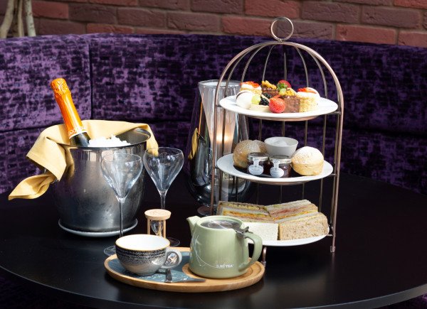 A table set with a champagne bucket, two empty flutes, a tiered stand of pastries and sandwiches, and teaware on a tray.