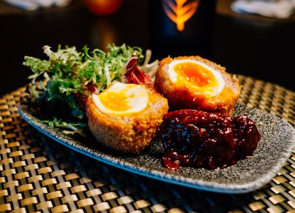 Two halved scotch eggs on a textured plate with mixed greens and a dark sauce.