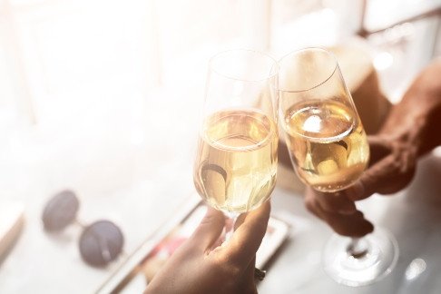 Two hands holding champagne flutes filled with a pale sparkling drink, raised in a toast near a bright window.