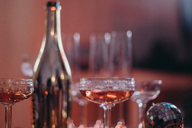 Several stemmed cocktail glasses filled with an amber drink sit beside a bottle on a table, with a disco ball in the foreground and blurred,