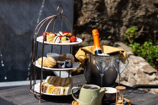 A tiered stand holding assorted pastries and sandwiches beside a bucket with a bottle of sparkling wine and two empty wine glasses on a dark