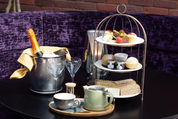 A table set with a bottle of champagne in an ice bucket, two empty glasses, a teapot and cup, and a tiered tray of sandwiches, pastries, and