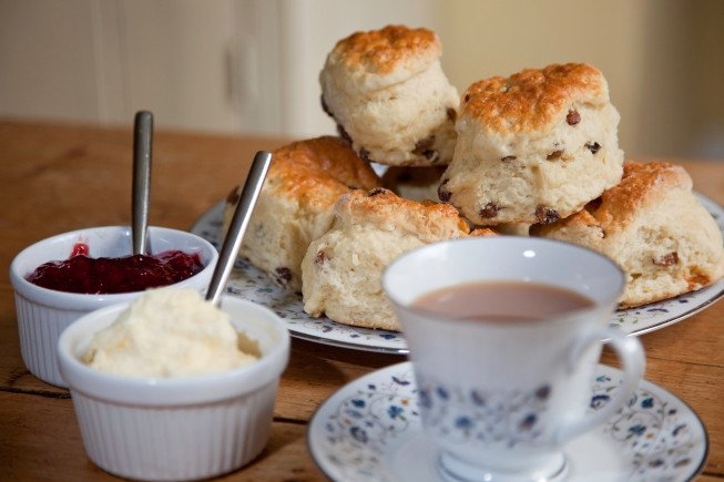 Plate of freshly baked scones with cups of jam, clotted cream, and tea on a wooden table.
