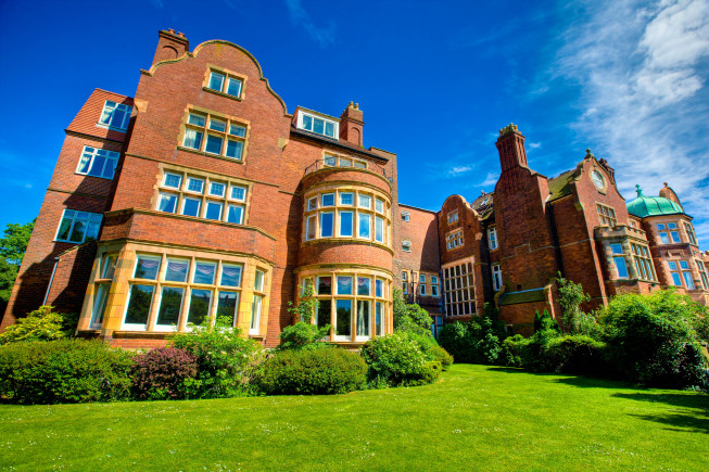 Large red-brick historic building with many windows, a curved central bay, and a green lawn in front under a blue sky.