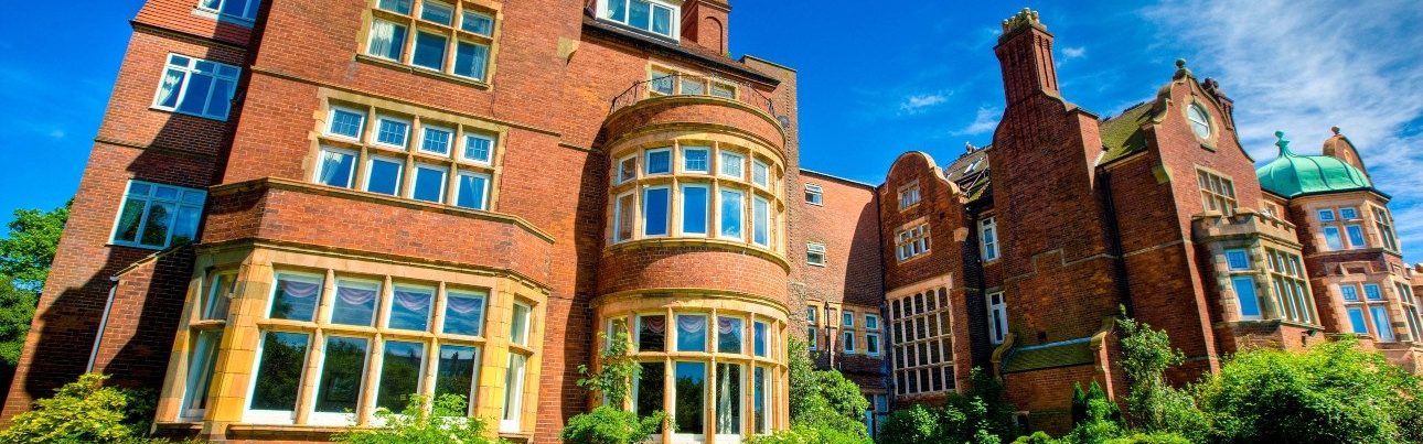 Wide view of a large historic brick building with many windows, rounded bay sections, and a green domed roof under a blue sky.