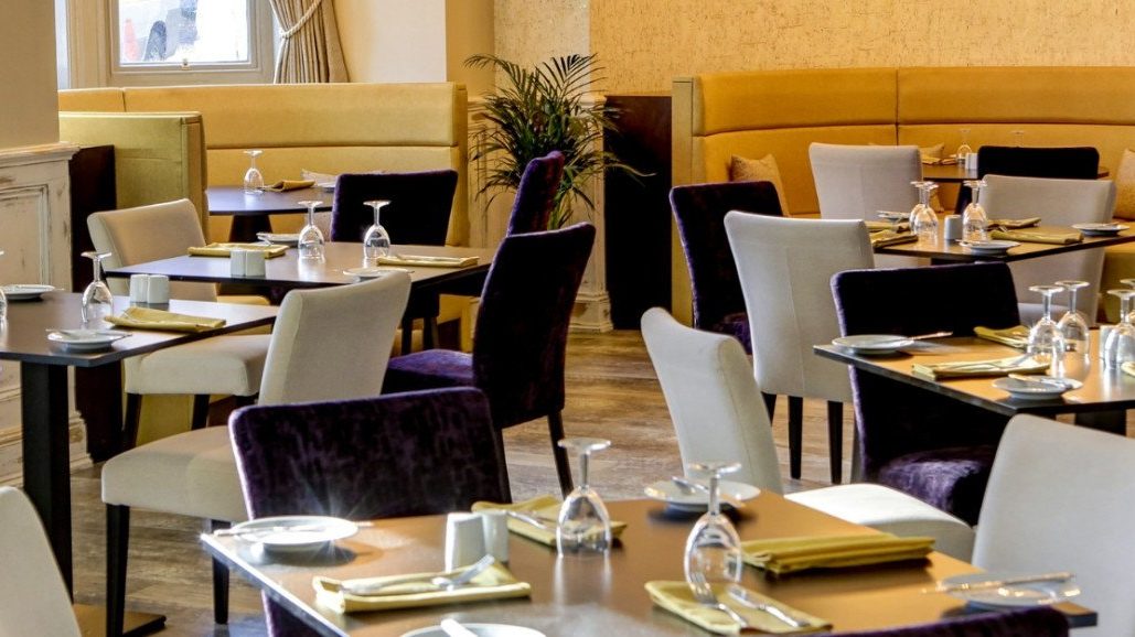 An empty restaurant dining room with set tables, upholstered chairs, and banquettes near tall windows.