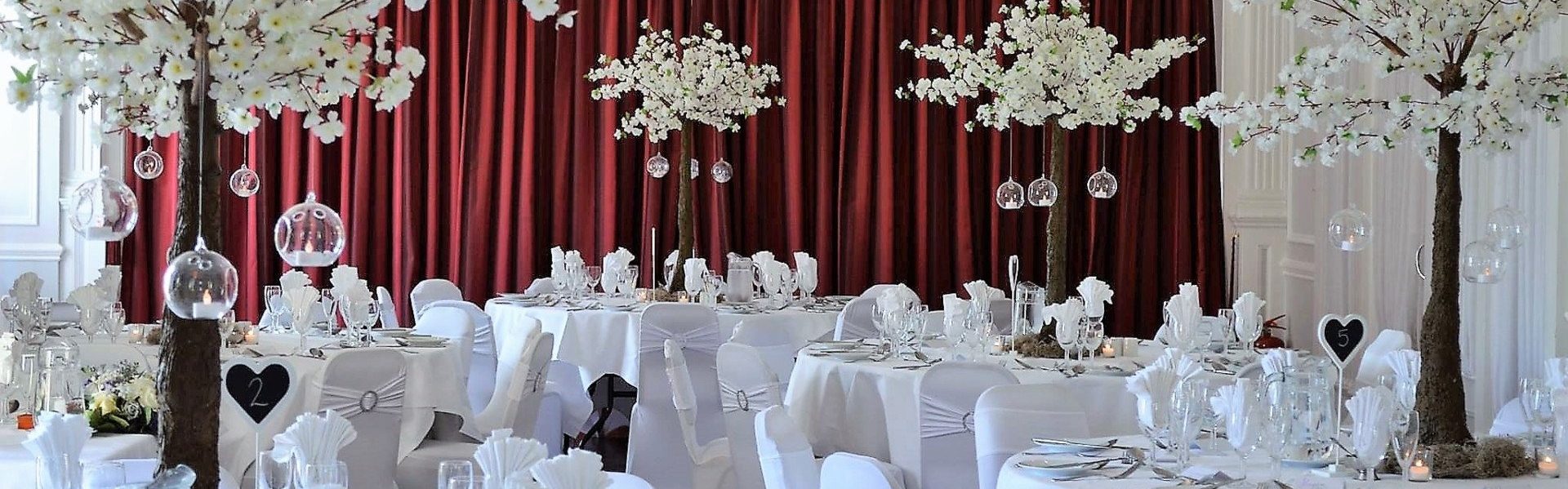 A banquet hall with round tables dressed in white tablecloths, white chair covers, and tall decorative trees with white blossoms and hanging