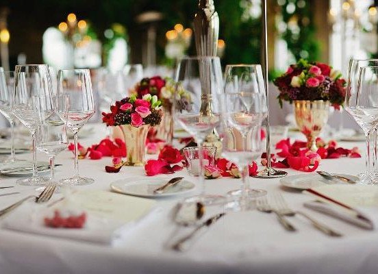 A banquet table set with wine glasses, plates, silverware, and small flower arrangements with pink rose petals scattered across a whitetable