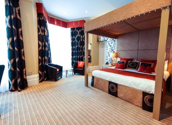 A bedroom with a large canopy bed, patterned curtains, two chairs by a bright window, and carpeted flooring.