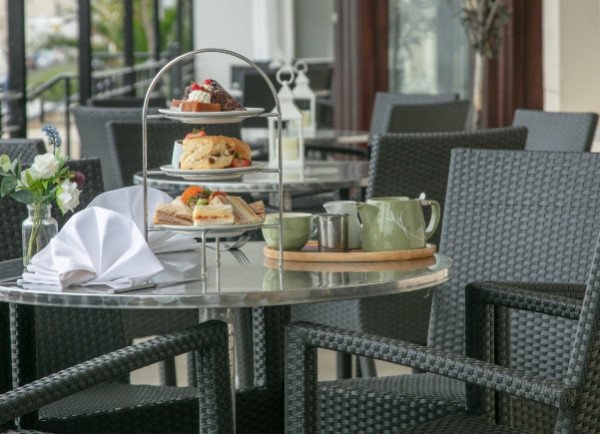A table on a patio with a tiered tray of pastries and sandwiches, a teapot, cups, a folded napkin, and a vase of flowers surrounded by woven