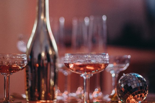 Glasses of amber-colored drink arranged beside a bottle, with a small disco ball in the foreground and blurred glassware in the background.