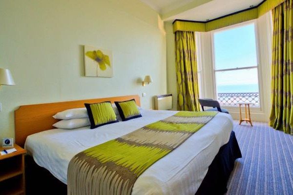 A neatly made hotel bed with green accents sits beside a large window with curtains, looking out toward the sea.