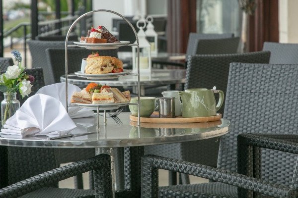 Tiered tray with pastries and sandwiches on a table with teapot, cups, and folded napkin in an outdoor seating area.