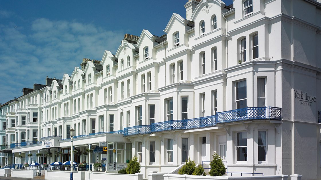 Row of white multi-story hotel buildings with blue balconies along a street under a blue sky.
