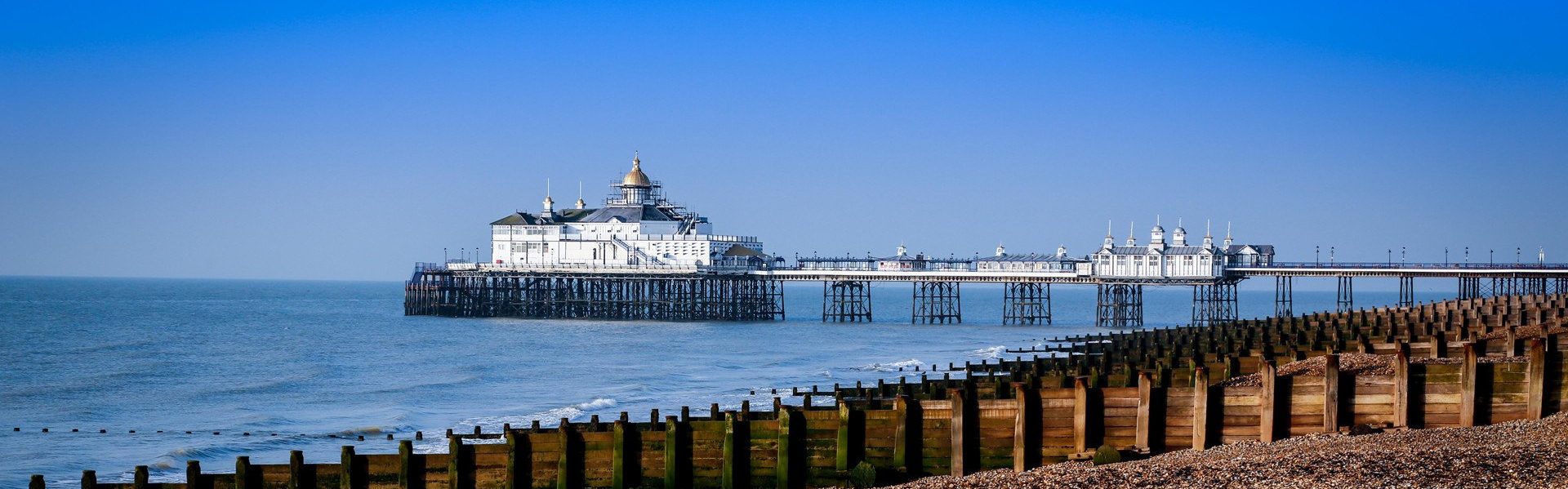 A long seaside pier with a large white building at the end stretches over calm blue water, with a pebbled shoreline and wooden groynes in a