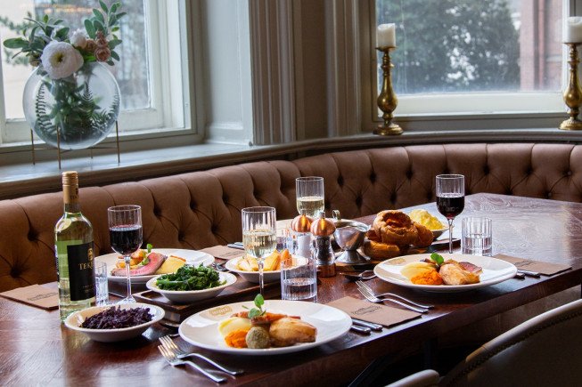 A set dining table with plated meals, wine glasses, water glasses, and a bottle of wine beside tufted booth seating near windows.