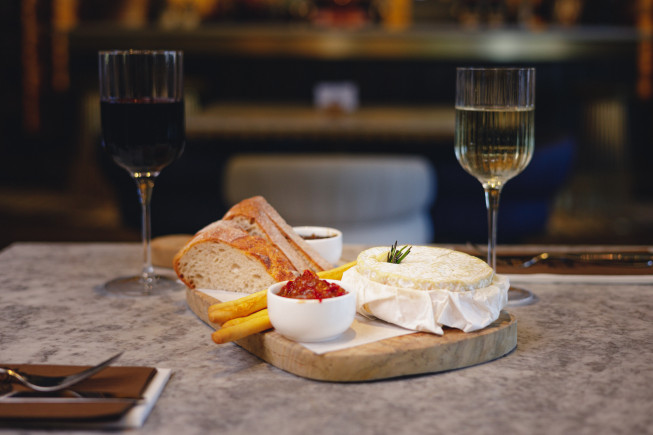 A wooden serving board with bread, cheese, condiments, and breadsticks sits between two glasses of wine on a table.