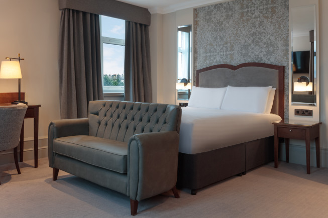 A hotel room with a neatly made bed, tufted sofa, desk, lamp, and window with curtains.