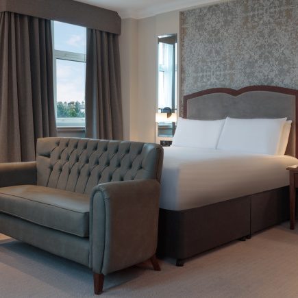 A hotel room with a neatly made bed, tufted sofa, desk, lamp, and window with curtains.