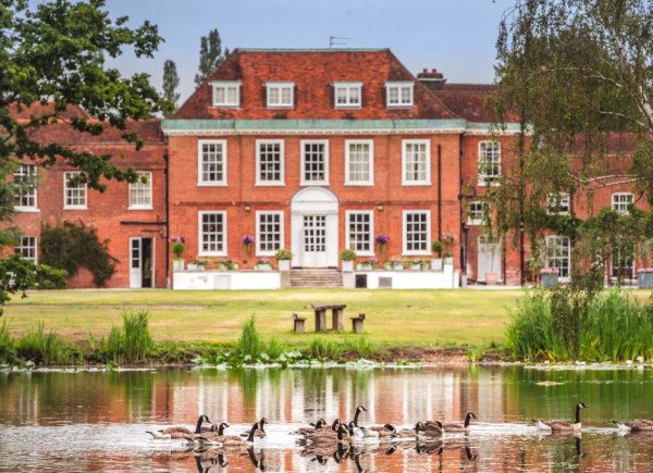 A red brick manor house with white-trimmed windows stands behind a grassy lawn and pond, with geese swimming in the foreground.
