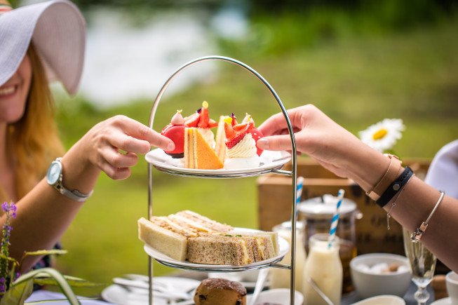 Two people reach toward a two-tiered tray of desserts and sandwiches at an outdoor table.