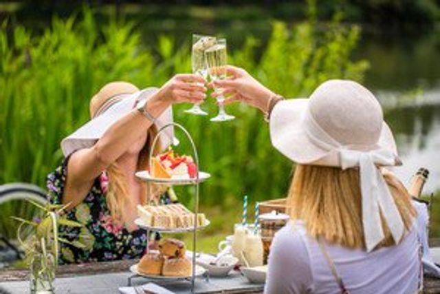 Two women in sun hats raise champagne flutes in a garden beside a tiered tea stand with sandwiches and pastries.